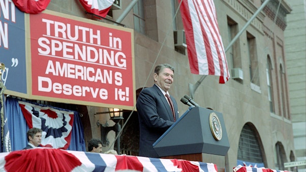 Reagan speaking on Truth in Spending American's Deserve It President Reagan speaking at a podium in front of brown building with red sign that reads "Truth in Spending American's Deserve It"