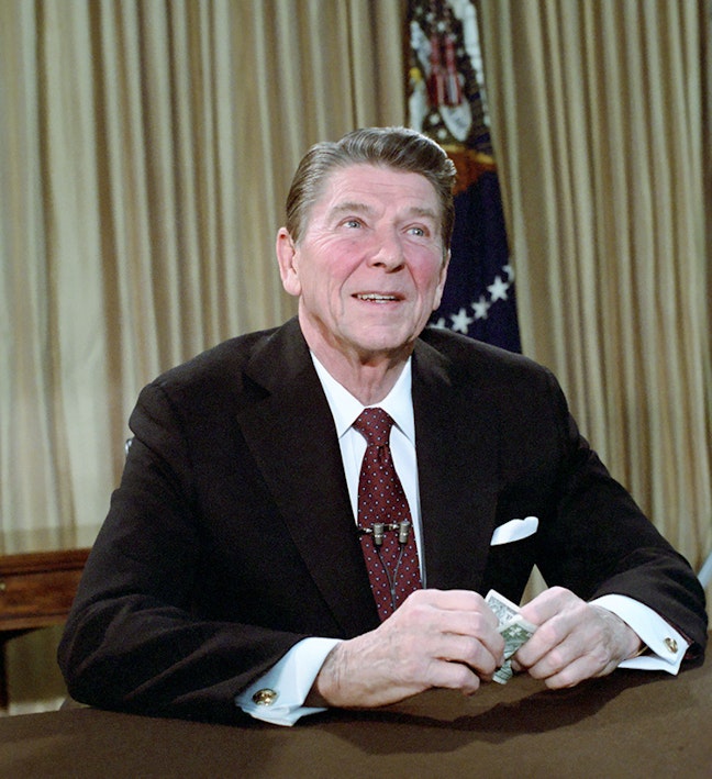President Reagan at desk holding one dollar bill Man sitting at desk holding one dollar bill